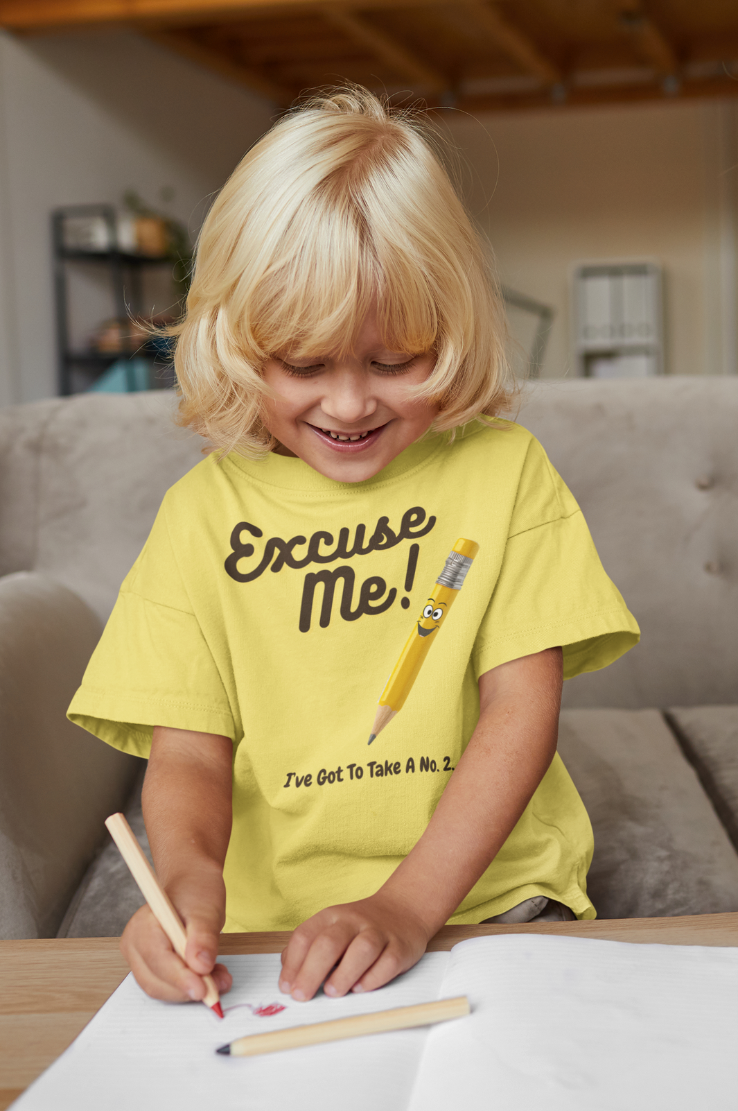 A little smiling boy writing on a desk wearing a t-shirt with the caption Excuse Me I've Got To Take A No.2. There is an illustrative smiling pencil alongside him