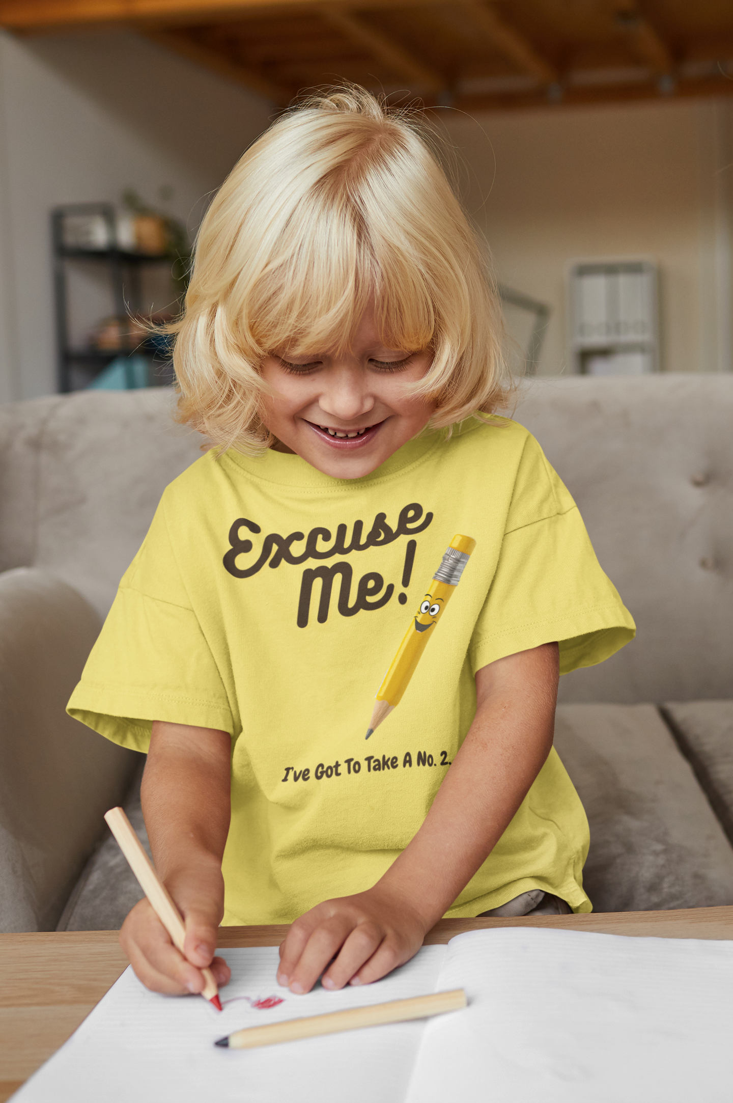 A little smiling boy writing on a desk wearing a t-shirt with the caption Excuse Me I've Got To Take A No.2. There is an illustrative smiling pencil alongside him